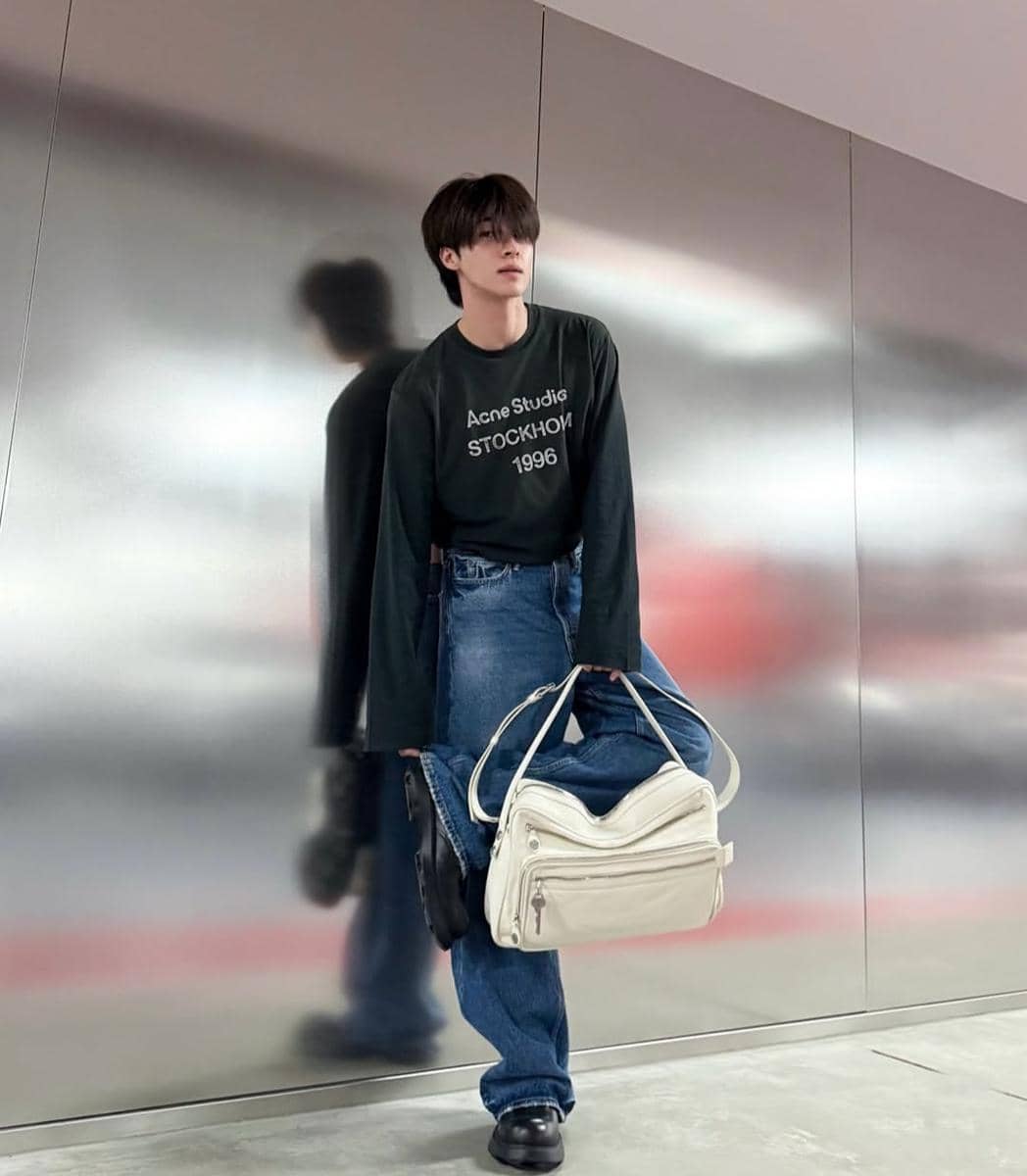 Seorang pria berpose di depan dinding logam mengenakan kaus hitam bertulisan, celana jeans longgar, sepatu hitam, dan tas bahu putih besar.