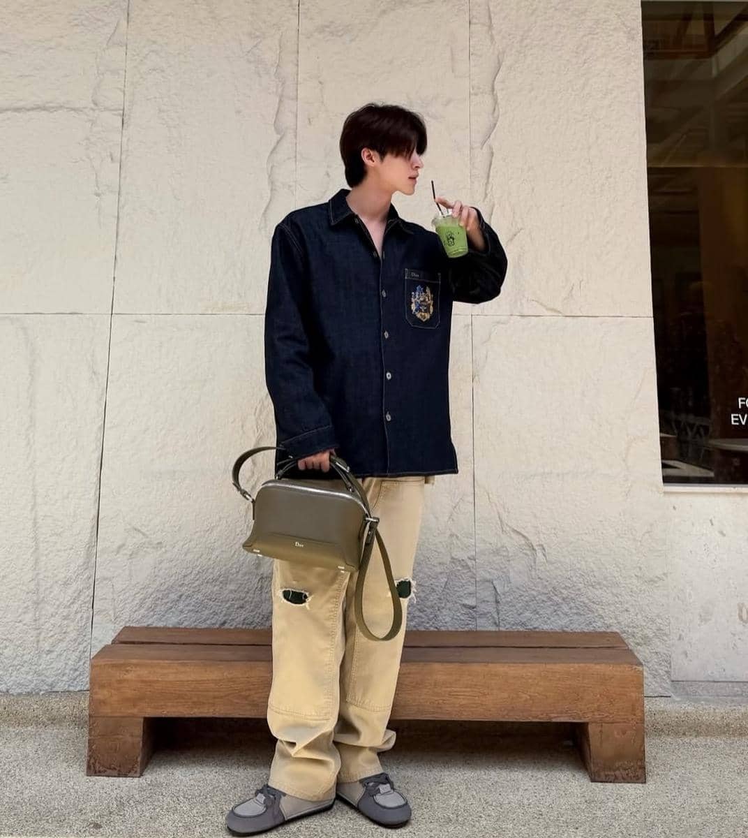 Seorang pria mengenakan kemeja denim biru tua dan celana krem sambil memegang tas hijau serta minum minuman hijau di depan dinding batu.