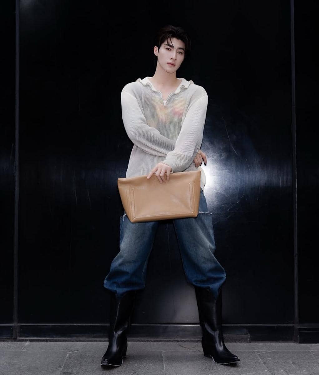 Seorang pria berpose dengan gaya kasual mengenakan sweater putih tipis, celana jeans longgar, sepatu bot hitam, dan memegang tas besar warna krem.