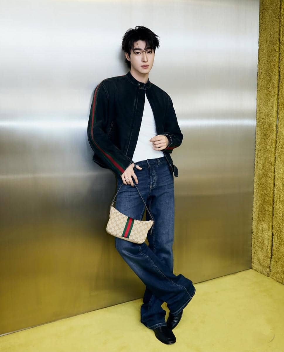 Seorang pria berpose santai mengenakan jaket hitam, kaus putih, dan celana jeans biru sambil memegang tas kecil bermotif di depan dinding metalik.