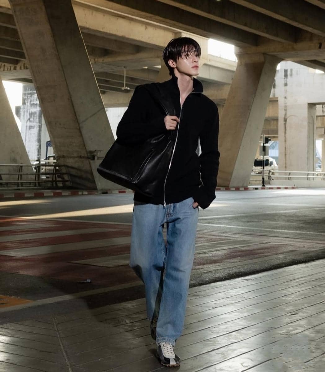 Seorang pria mengenakan jaket hitam, celana jeans longgar, dan sepatu kasual berjalan di bawah jembatan beton sambil membawa tas besar hitam.
