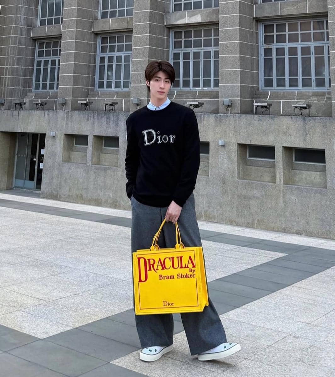 Seorang pria bergaya kasual mengenakan sweater hitam bertuliskan Dior, celana abu-abu longgar, dan membawa tas kuning besar di area luar gedung.