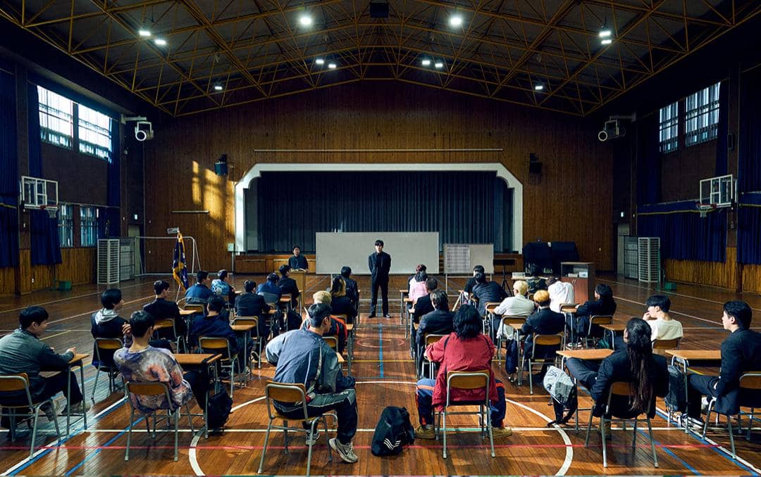 Suasana kelas di aula sekolah dengan banyak orang duduk di kursi menghadap panggung, diambil dari drama Korea Teach You a Lesson.