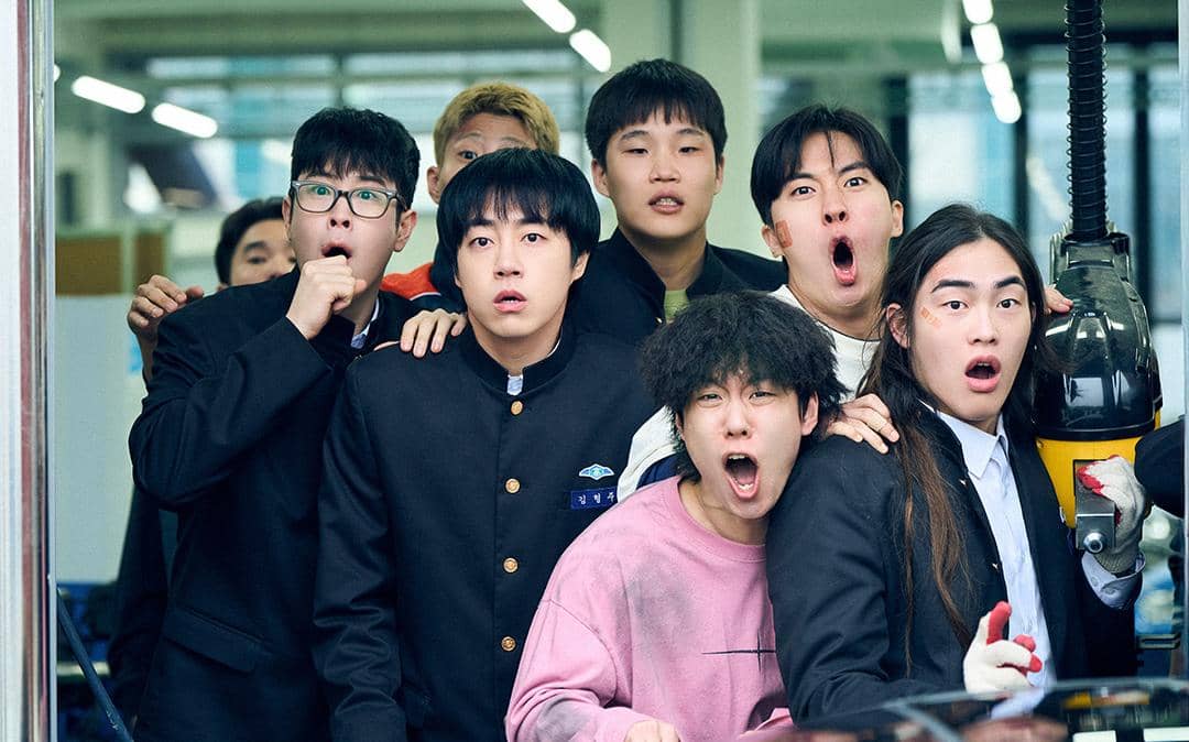Sekelompok siswa laki-laki dengan ekspresi terkejut dan penasaran berdiri berdekatan di dalam ruang sekolah dalam adegan drama Korea Teach You a Lesson.