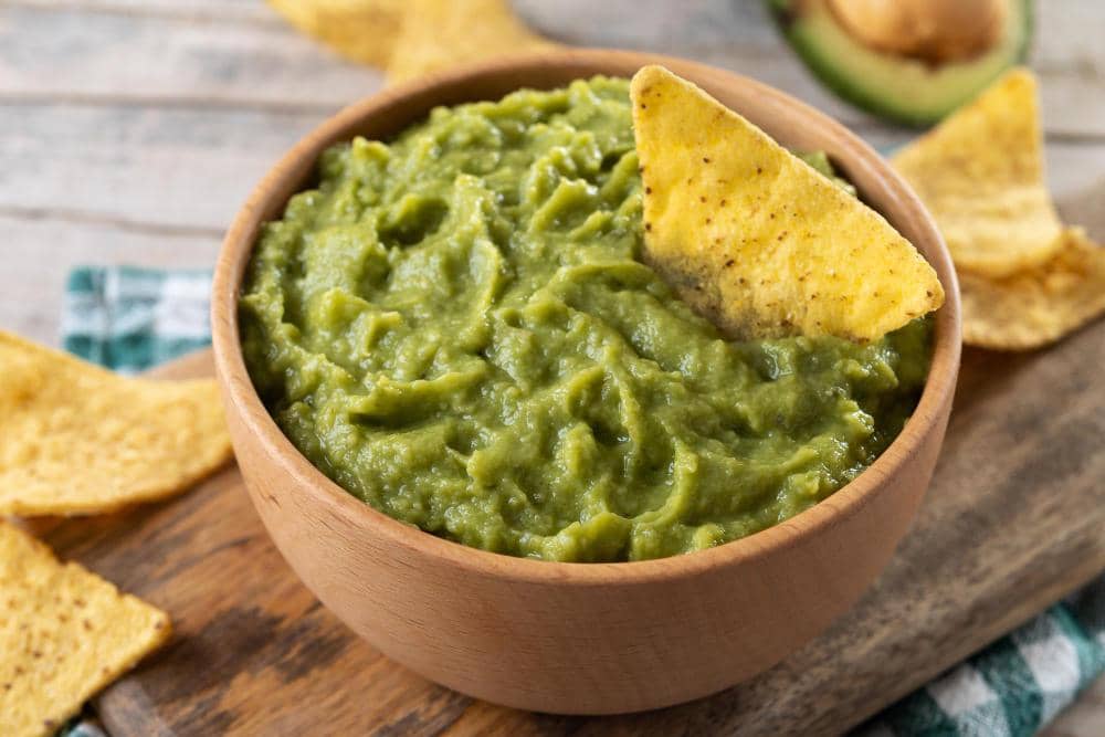 Semangkuk guacamole hijau kental disajikan dalam mangkuk kayu dengan keripik tortilla di atasnya di atas meja kayu.