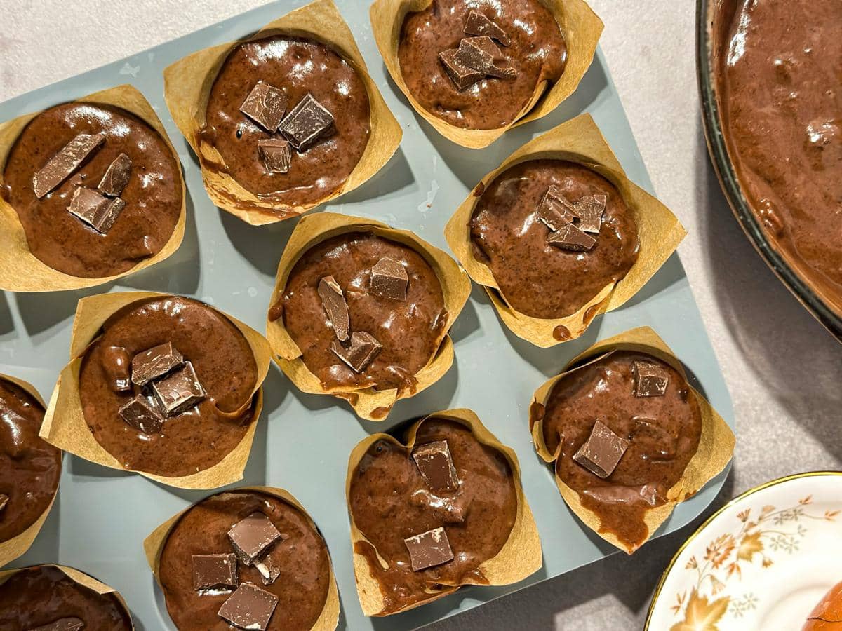 Adonan muffin cokelat dalam cetakan kertas dengan potongan cokelat di atasnya siap untuk dipanggang di atas meja dapur.