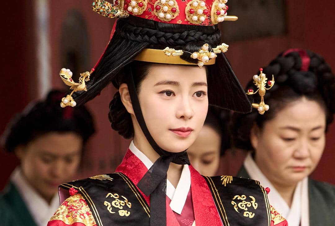 Aktris mengenakan hanbok kerajaan berwarna merah dan hitam dengan hiasan kepala tradisional dalam adegan drama Korea My Royal Nemesis.
