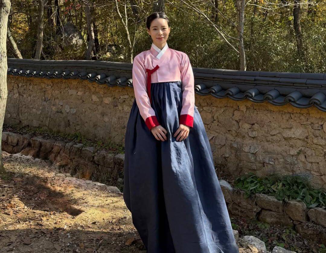 Seorang wanita mengenakan hanbok tradisional Korea berwarna merah muda dan biru tua berdiri di depan dinding batu di area taman yang rindang.