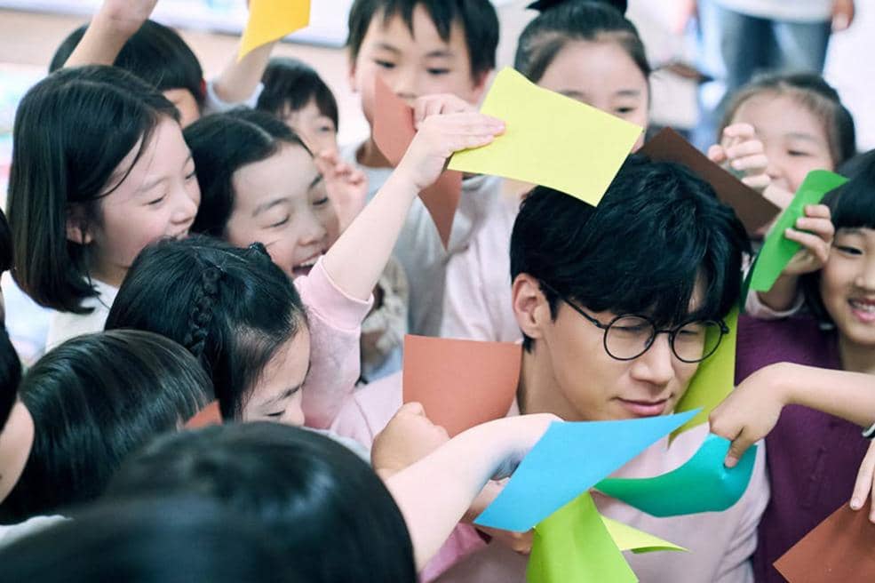 Seorang pria berkacamata dikelilingi anak-anak yang memegang kertas warna-warni dalam suasana ceria di ruang kelas.
