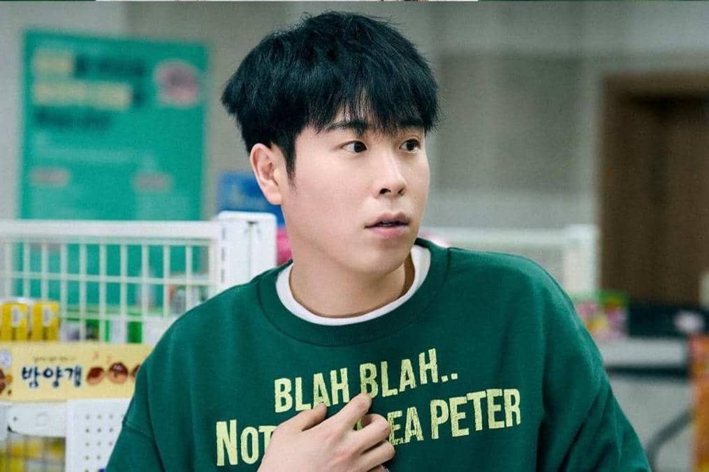 Seorang pria berambut pendek mengenakan sweater hijau dengan tulisan kuning sedang berdiri di dalam ruangan seperti toko atau minimarket.
