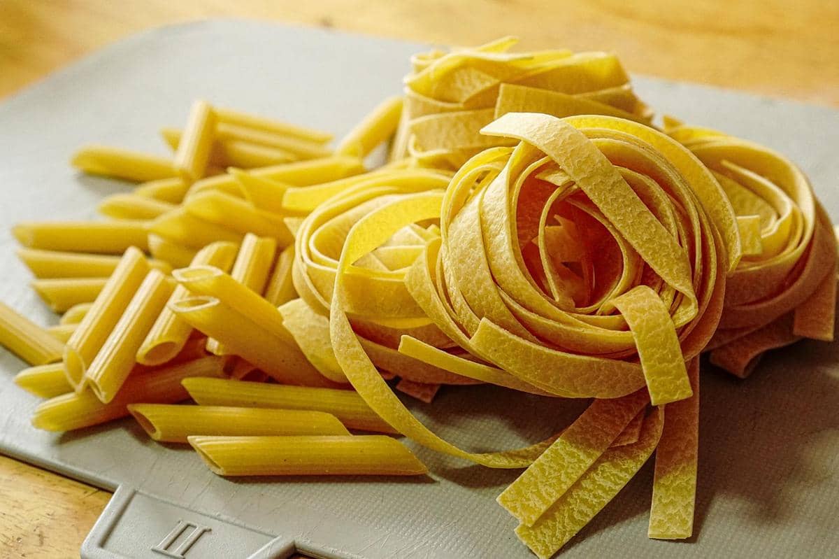 Tumpukan pasta kering jenis fettuccine dan penne berwarna kuning keemasan di atas permukaan dapur dengan pencahayaan hangat.