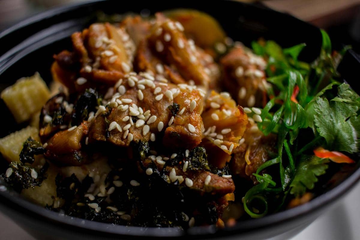 Semangkuk beef bulgogi dengan taburan biji wijen, potongan daging sapi berbumbu, sayuran hijau, dan nasi di dalam mangkuk hitam.