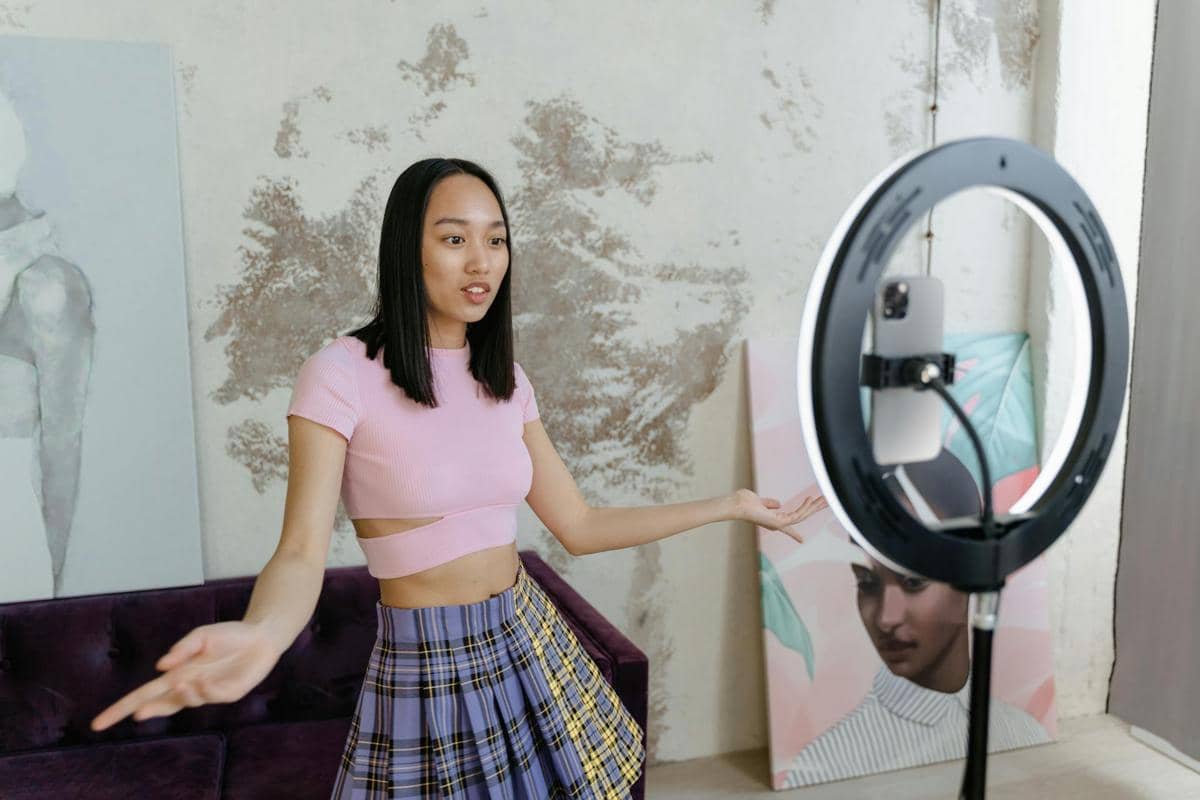 Seorang perempuan mengenakan atasan pink dan rok kotak-kotak sedang melakukan siaran langsung di media sosial menggunakan ponsel dan ring light.