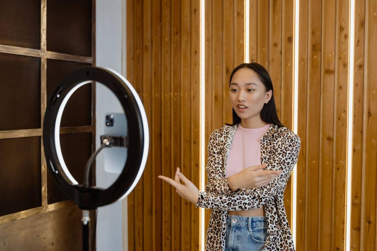 Seorang perempuan mengenakan atasan bermotif leopard sedang berbicara di depan kamera ponsel dengan lampu ring light di ruangan berpapan kayu.