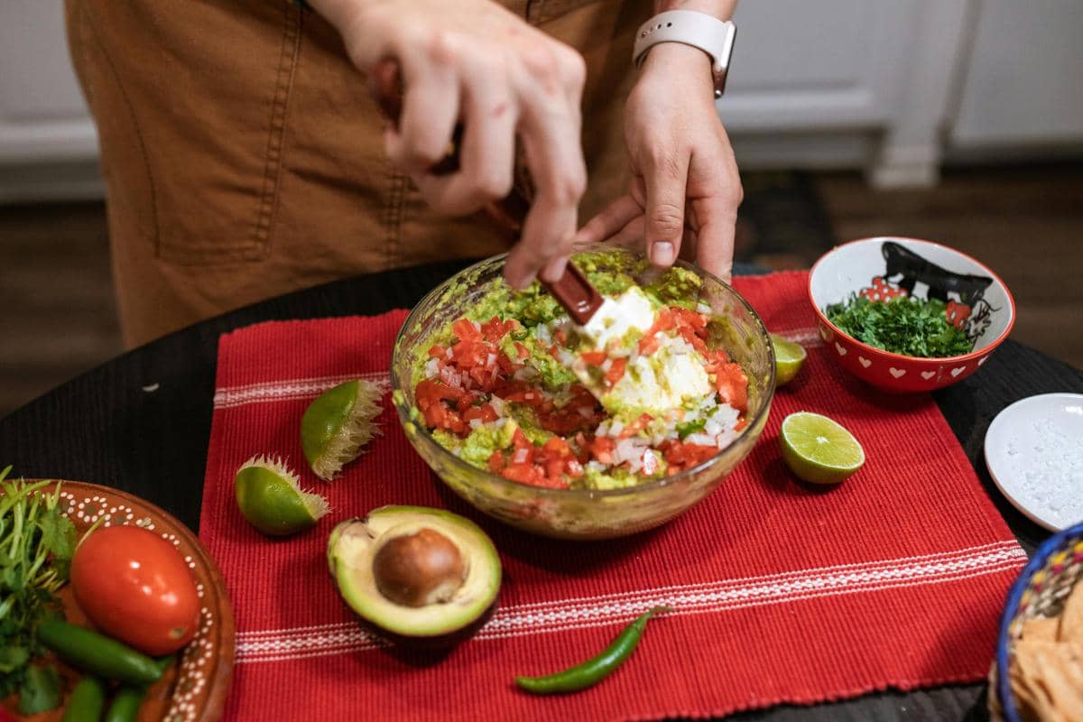 Seseorang sedang mencampur bahan guacamole seperti alpukat, tomat, bawang, dan jeruk nipis di atas meja dengan alas kain merah.