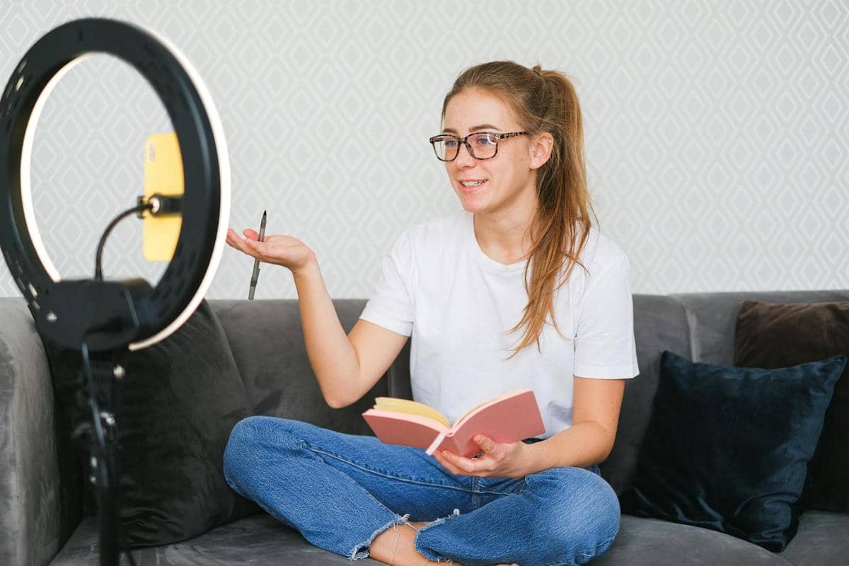 Seorang perempuan duduk di sofa sambil berbicara di depan kamera ponsel dengan lampu ring light, memegang buku catatan dan pena.
