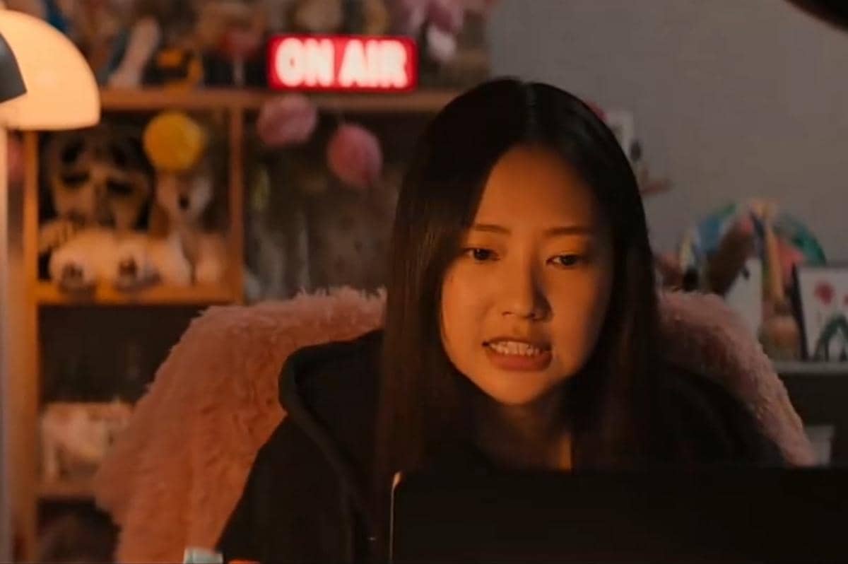 Seorang perempuan muda duduk di depan laptop dengan ekspresi tegang di kamar yang diterangi lampu hangat dan tanda 'ON AIR' menyala di belakangnya.
