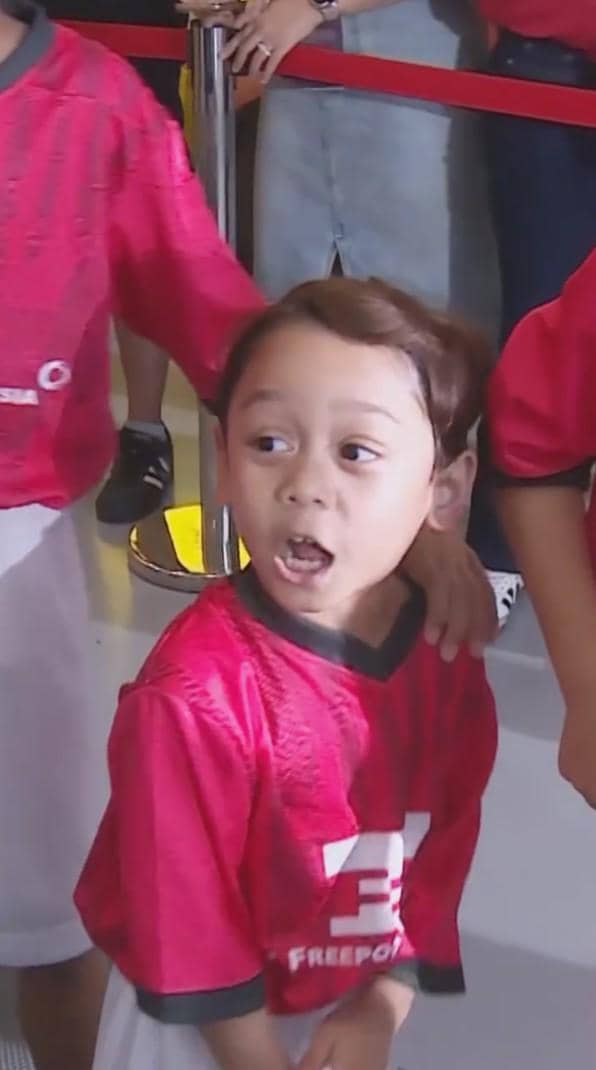 Seorang anak mengenakan jersey merah tim nasional berdiri di area publik dengan ekspresi antusias sambil dikelilingi anak-anak lain.