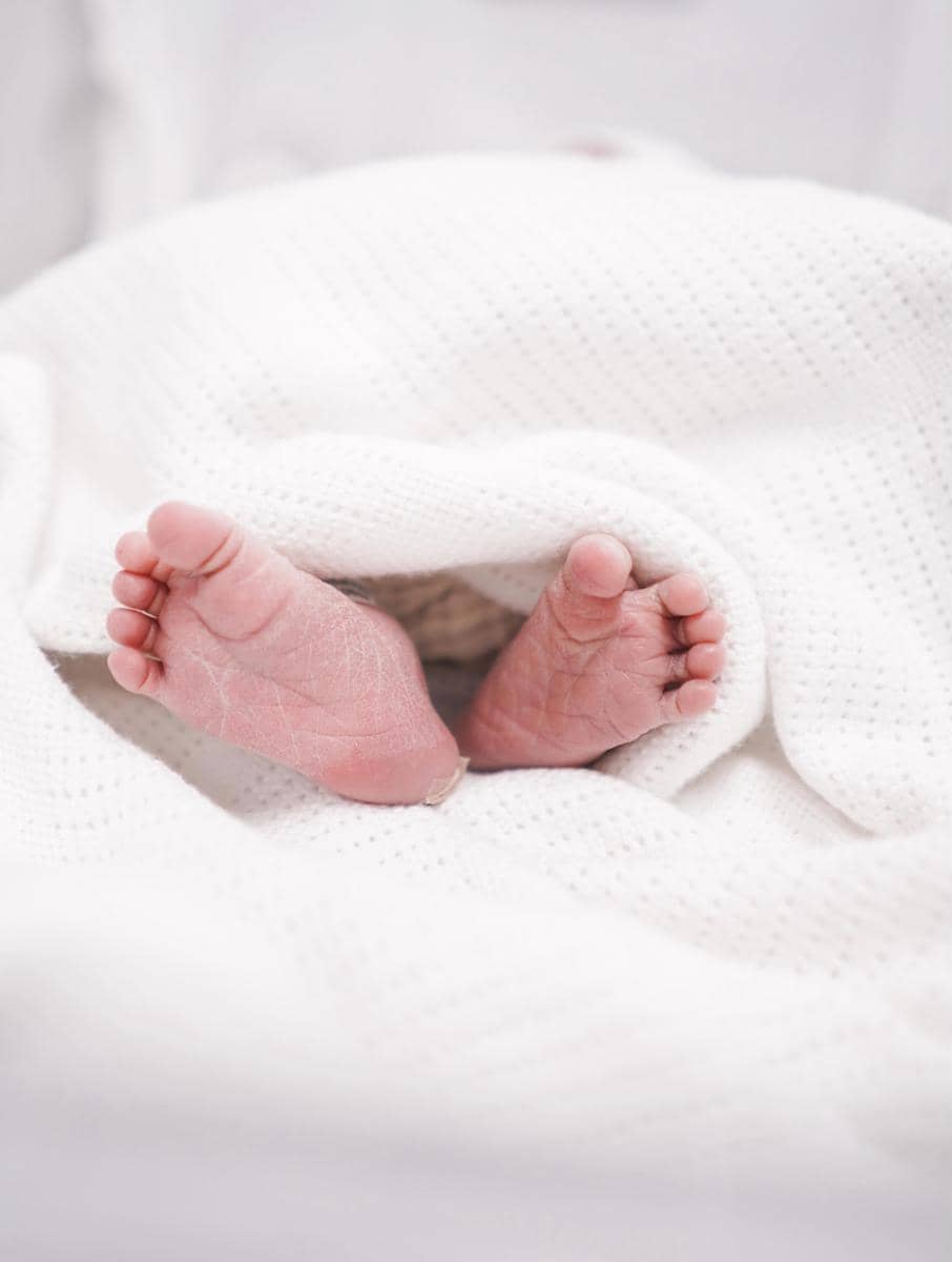 Kaki bayi yang baru lahir tampak mungil dan lembut, diselimuti kain putih berlubang halus di atas permukaan tempat tidur.