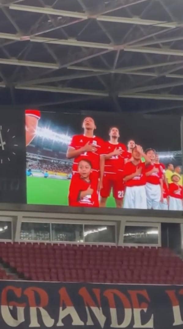 Layar stadion menampilkan pemain tim sepak bola mengenakan seragam merah bersama anak-anak pendamping sebelum pertandingan dimulai.
