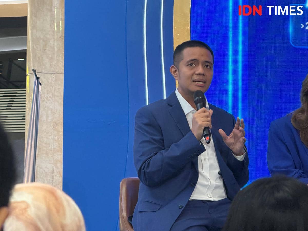 Seorang pria mengenakan setelan jas biru sedang berbicara sambil memegang mikrofon di depan audiens dalam sebuah acara.
