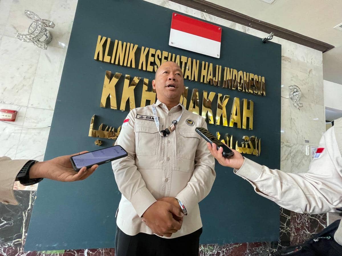 Seorang pejabat Kementerian Haji Indonesia mengenakan seragam krem sedang memberikan keterangan kepada wartawan di depan papan Klinik Kesehatan Haji Indonesia Makkah.