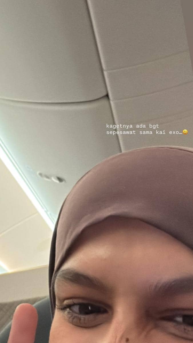 Perempuan berhijab tersenyum sambil berpose di dalam kabin pesawat dengan latar langit-langit pesawat berwarna terang.