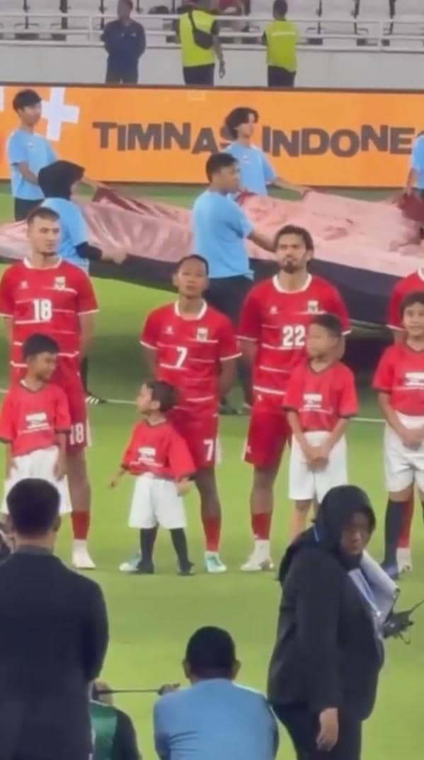 Anak kecil mengenakan kaus merah berdiri di depan pemain timnas Indonesia yang berbaris di lapangan sebelum pertandingan dimulai.