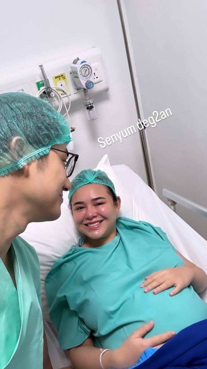 Seorang wanita hamil tersenyum di ranjang rumah sakit mengenakan pakaian operasi hijau, didampingi pria yang juga memakai pakaian medis.