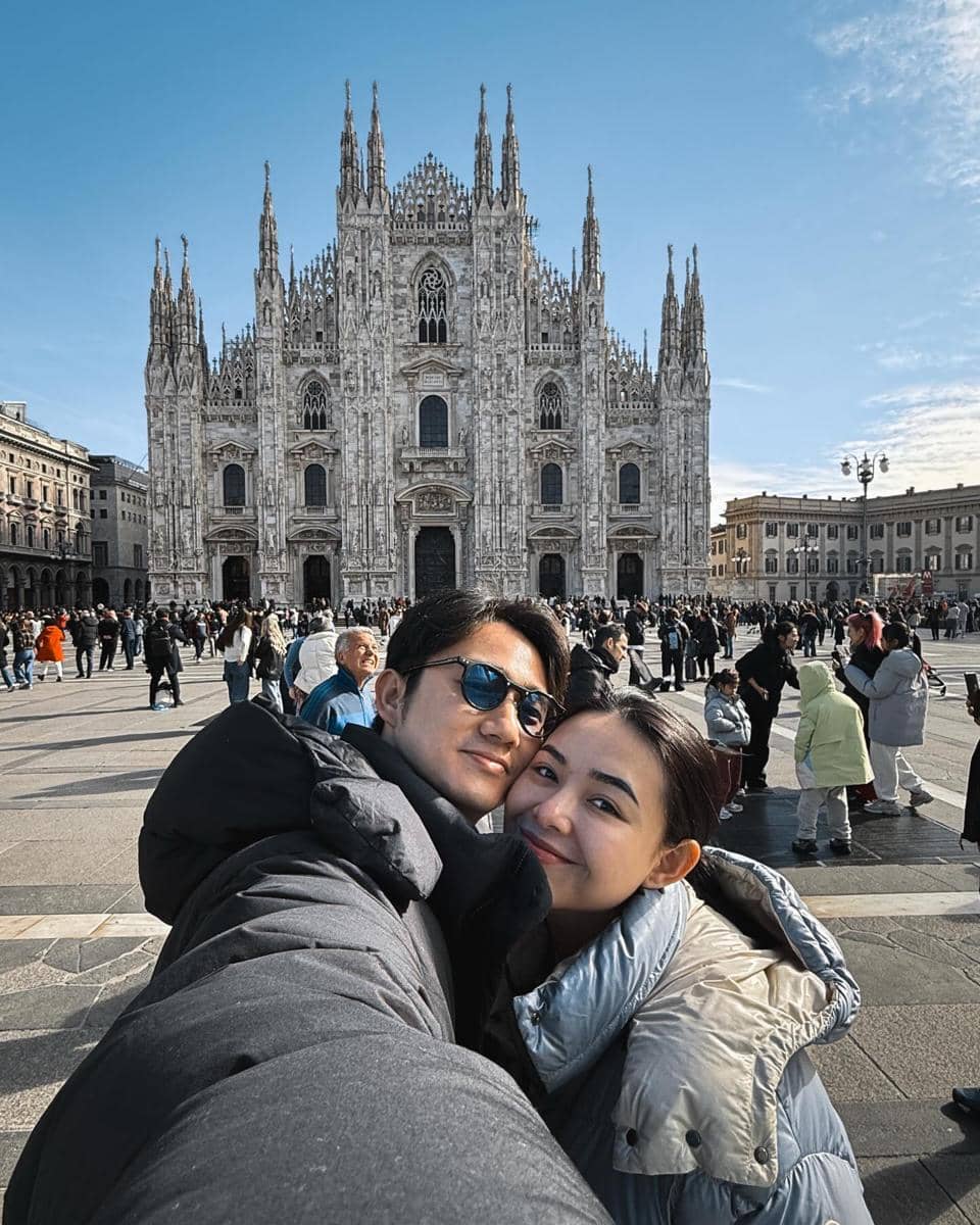 Sepasang kekasih berpose bersama di depan Katedral Milan, Italia, dengan latar langit cerah dan keramaian wisatawan di alun-alun.