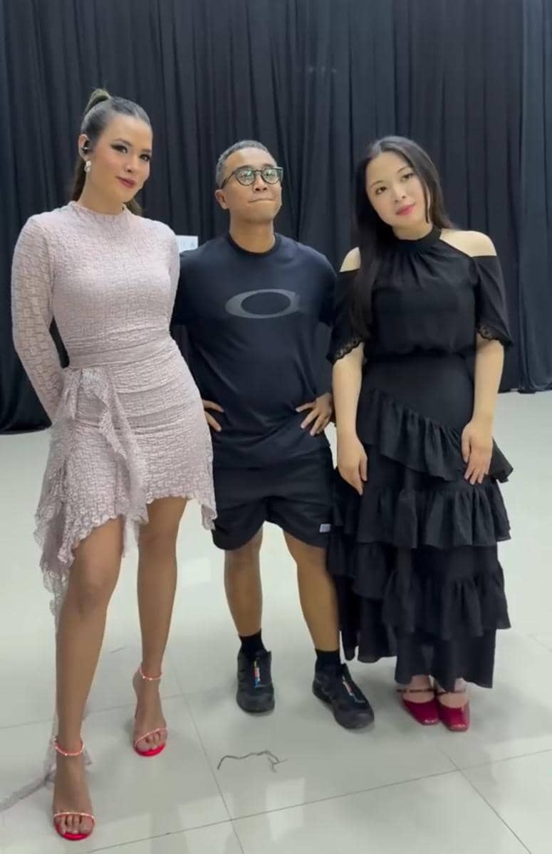 Tiga orang berpose bersama di dalam ruangan dengan latar tirai hitam, dua wanita mengenakan gaun elegan dan satu pria memakai pakaian kasual.