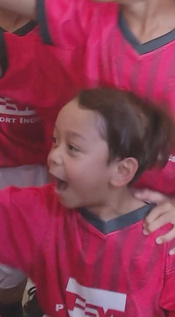 Seorang anak mengenakan jersey merah tim sepak bola dengan ekspresi gembira di antara teman-temannya yang juga memakai seragam serupa.