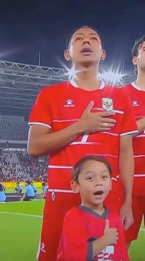 Seorang pemain timnas Indonesia berdiri dengan tangan di dada bersama seorang anak kecil mengenakan jersey merah di stadion.