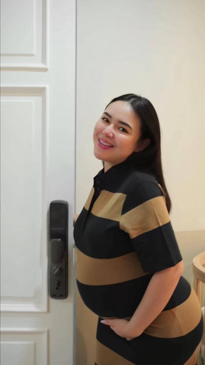 Seorang wanita hamil tersenyum mengenakan gaun bergaris hitam dan cokelat sambil berdiri di depan pintu berwarna putih.