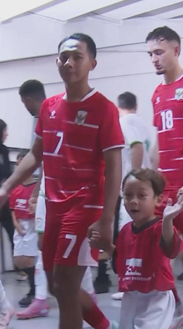 Seorang pemain timnas Indonesia mengenakan seragam merah berjalan di lorong stadion sambil menggandeng anak kecil pendamping pemain.