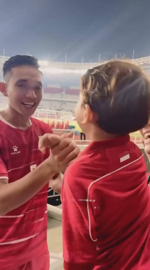 Seorang pemain sepak bola timnas Indonesia tersenyum dan berjabat tangan dengan anak kecil yang mengenakan jersey merah di stadion.