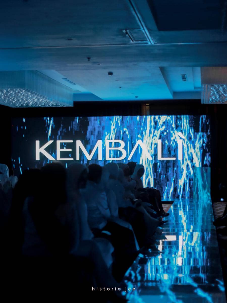 Suasana fashion show dengan pencahayaan biru dan layar besar bertuliskan 'KEMBALI' di ujung panggung, dihadiri para penonton.