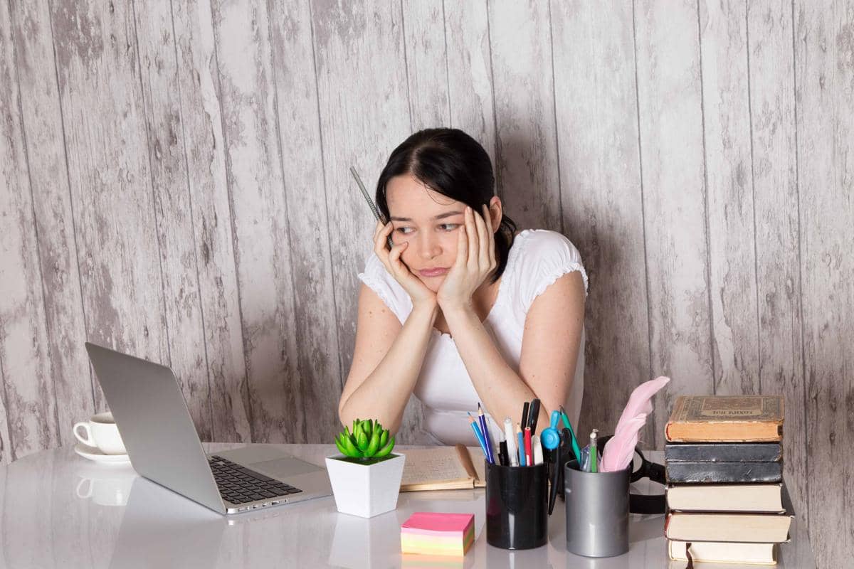 Seorang wanita duduk di meja kerja dengan ekspresi sedih sambil menatap laptop, dikelilingi buku dan alat tulis.