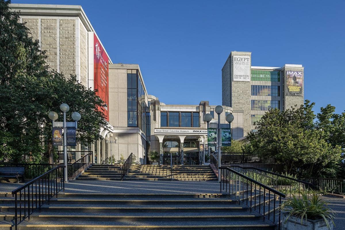 Tampak depan Royal BC Museum di Victoria, Kanada, dengan tangga menuju pintu masuk dan gedung modern berlatar langit biru cerah.