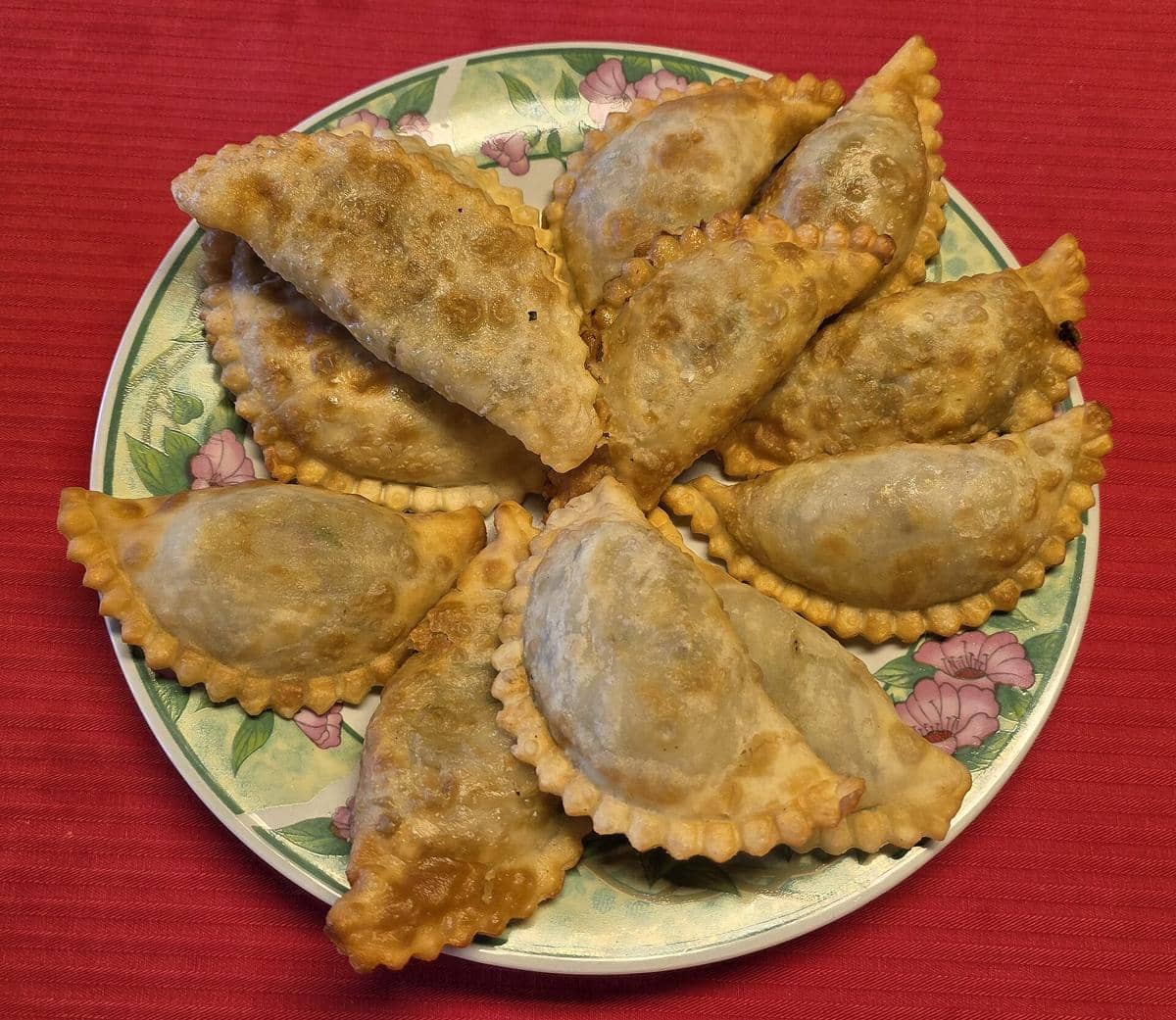 Beberapa empanada goreng berwarna keemasan tersusun di atas piring bermotif bunga dengan alas kain merah.