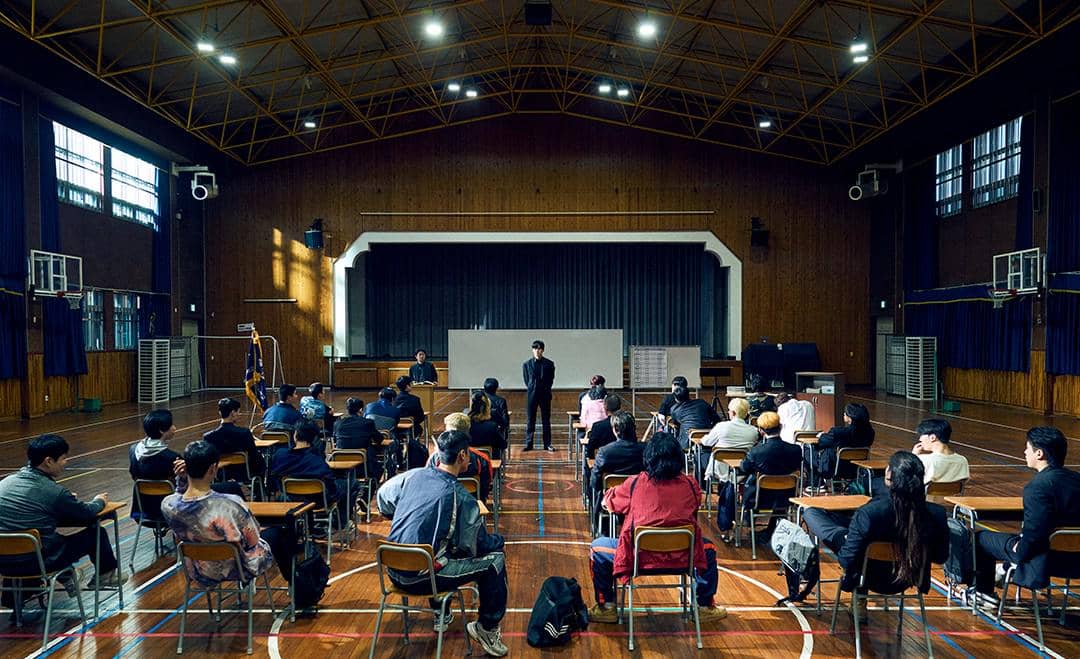 Sejumlah orang duduk di kursi menghadap panggung di aula sekolah dengan pencahayaan alami dari jendela besar dan langit-langit tinggi.