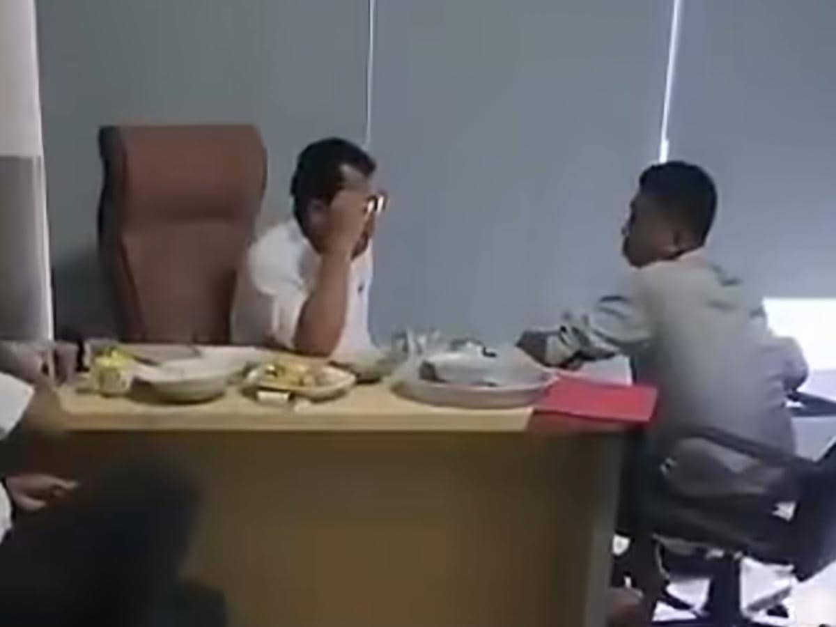 Dua pria duduk di ruang kerja dengan meja berisi makanan dan dokumen, salah satunya tampak berbicara sambil memegang kepala.