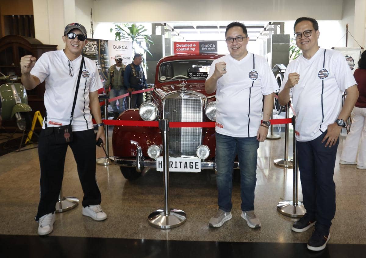 Tiga pria berpose di depan mobil klasik berwarna merah yang dipamerkan di sebuah acara otomotif dalam ruangan.