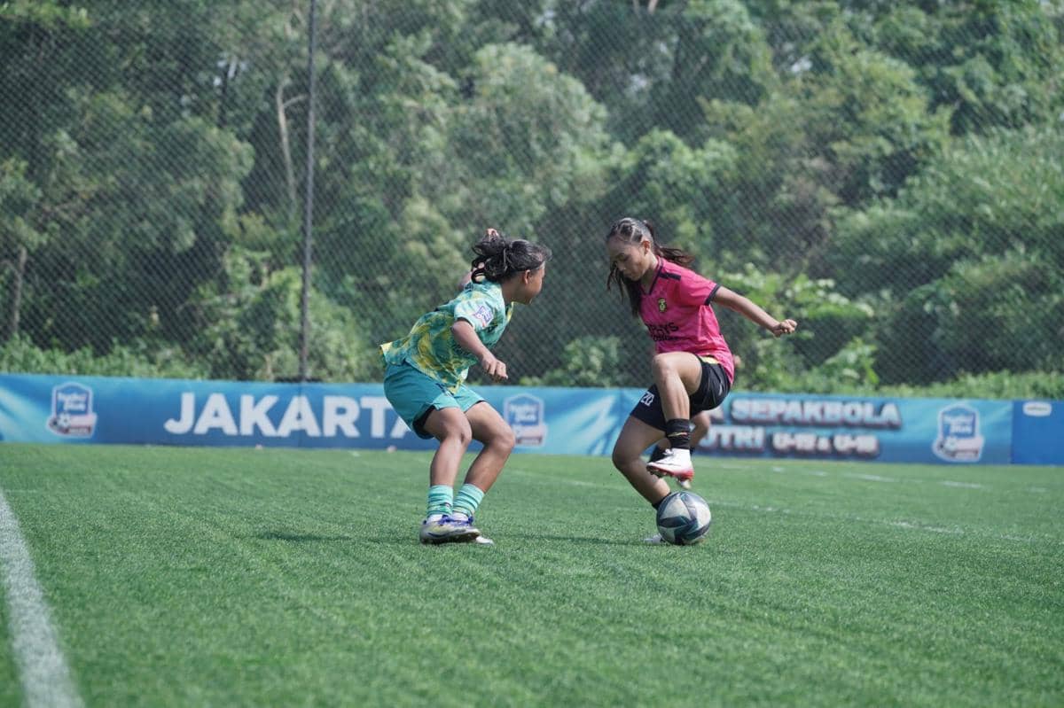 Dua pemain muda perempuan bertanding di lapangan sepak bola dalam ajang Hydroplus Soccer League Jakarta dengan latar pepohonan hijau.