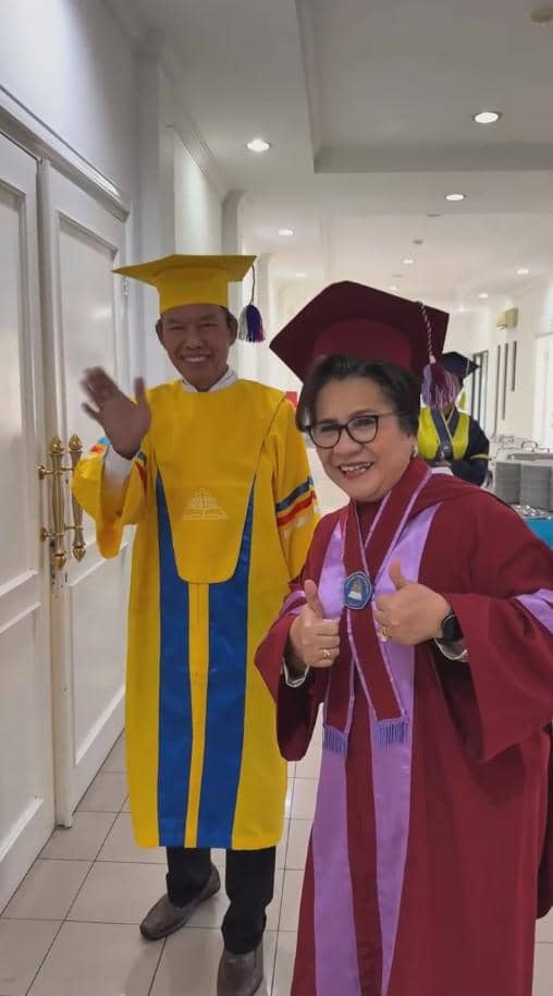 Dua wisudawan lanjut usia mengenakan toga berwarna kuning dan merah marun tersenyum bahagia di lorong gedung wisuda.