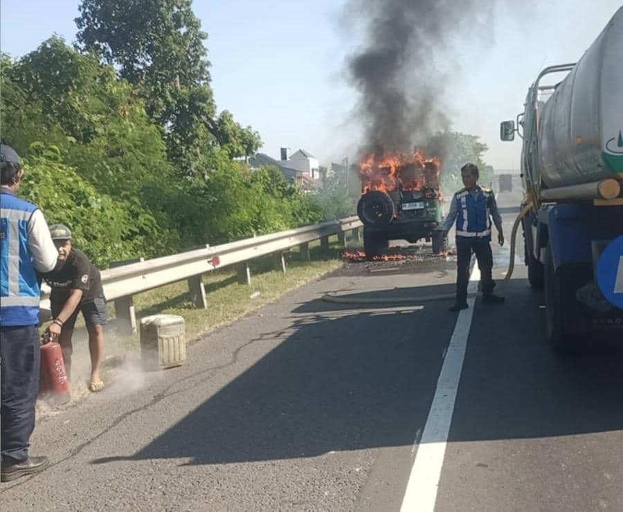 Mobil Toyota Land Cruiser tahun 1973 terbakar di Jalan Tol Palikanci arah Jakarta dengan asap hitam tebal dan petugas berusaha memadamkan api.