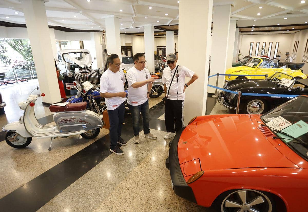 Tiga pria berbaju putih berbincang di antara deretan mobil dan motor klasik dalam sebuah pameran kendaraan di dalam ruangan.