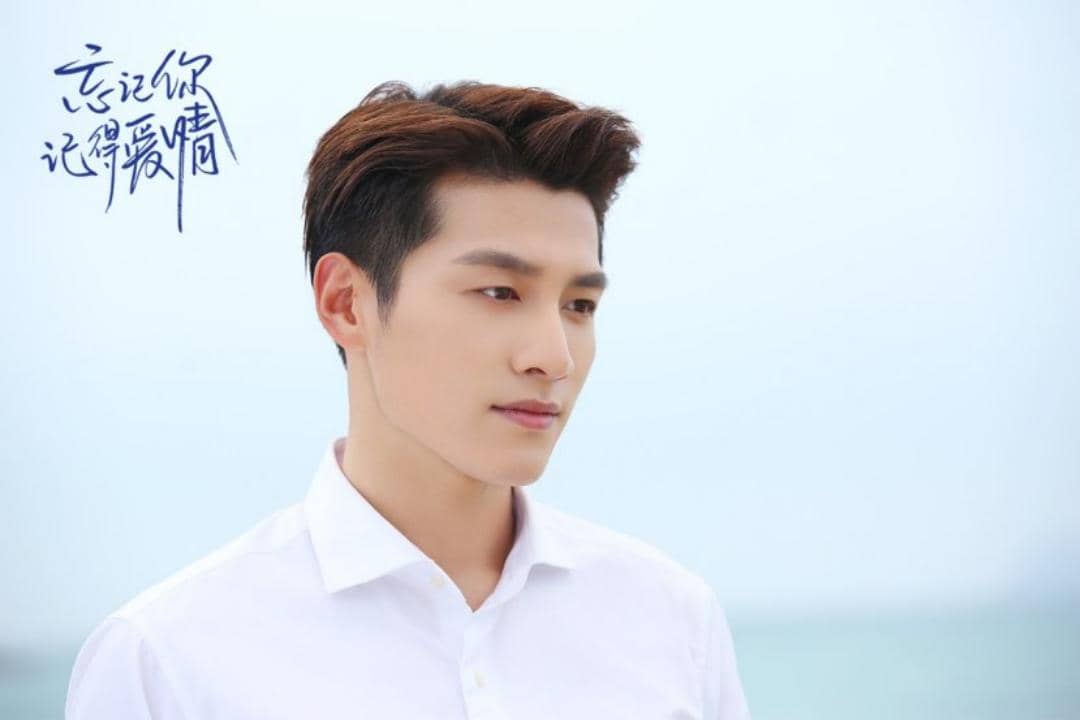 Seorang pria berambut pendek mengenakan kemeja putih berdiri di tepi pantai dengan latar langit cerah dalam adegan drama Forget You Remember Love.