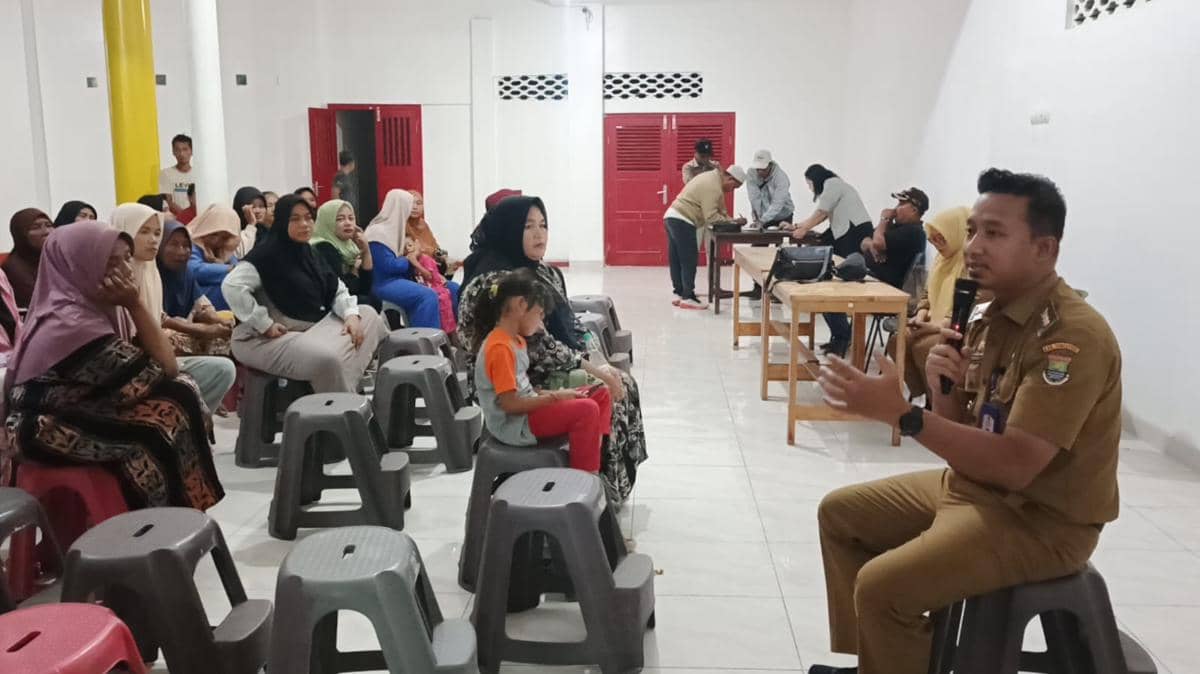 Seorang pejabat kecamatan berbicara menggunakan mikrofon di depan warga yang duduk di kursi plastik dalam sebuah pertemuan di ruangan tertutup.