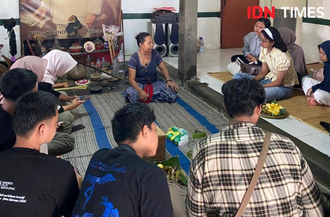Seorang perempuan berbicara di hadapan sekelompok anak muda yang duduk melingkar di ruangan tradisional dengan alat musik dan makanan di sekitarnya.